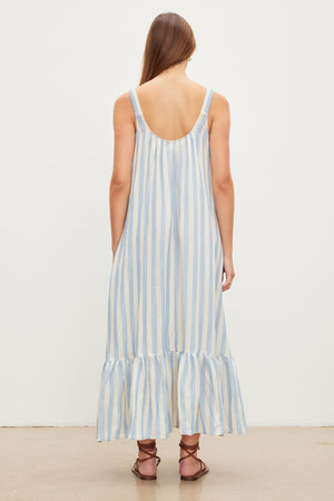 A person with long brown hair is wearing the Velvet by Graham & Spencer MERADITH STRIPED LINEN MAXI DRESS, which is sleeveless, blue and white striped, maxi length with a low back and ruffled hem, standing against a plain white background.