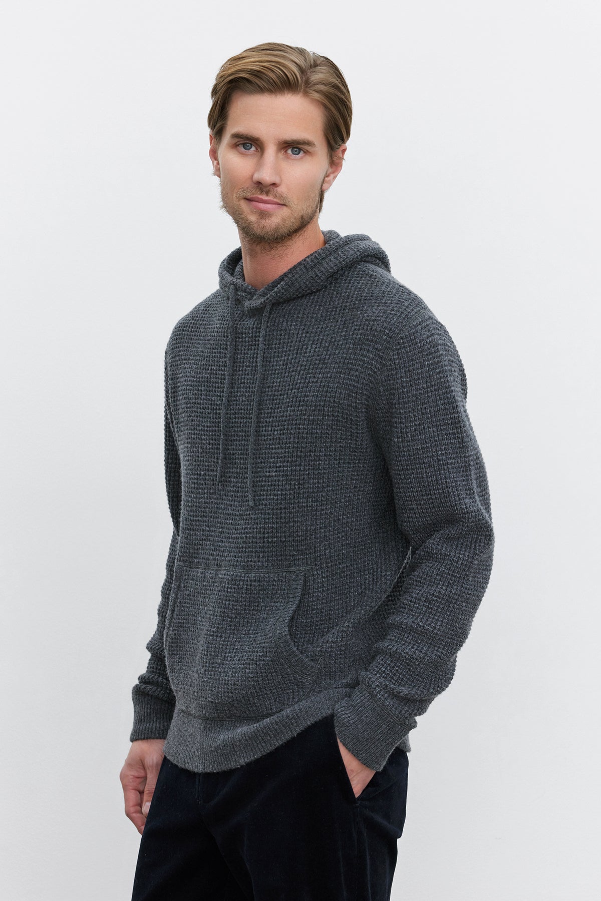 A man is standing against a plain light background, wearing the Velvet by Graham & Spencer SHANE SWEATER HOODIE. With his hands in the front pockets of the gray textured pullover, he exudes an aura of warmth and comfort.-37893929763009