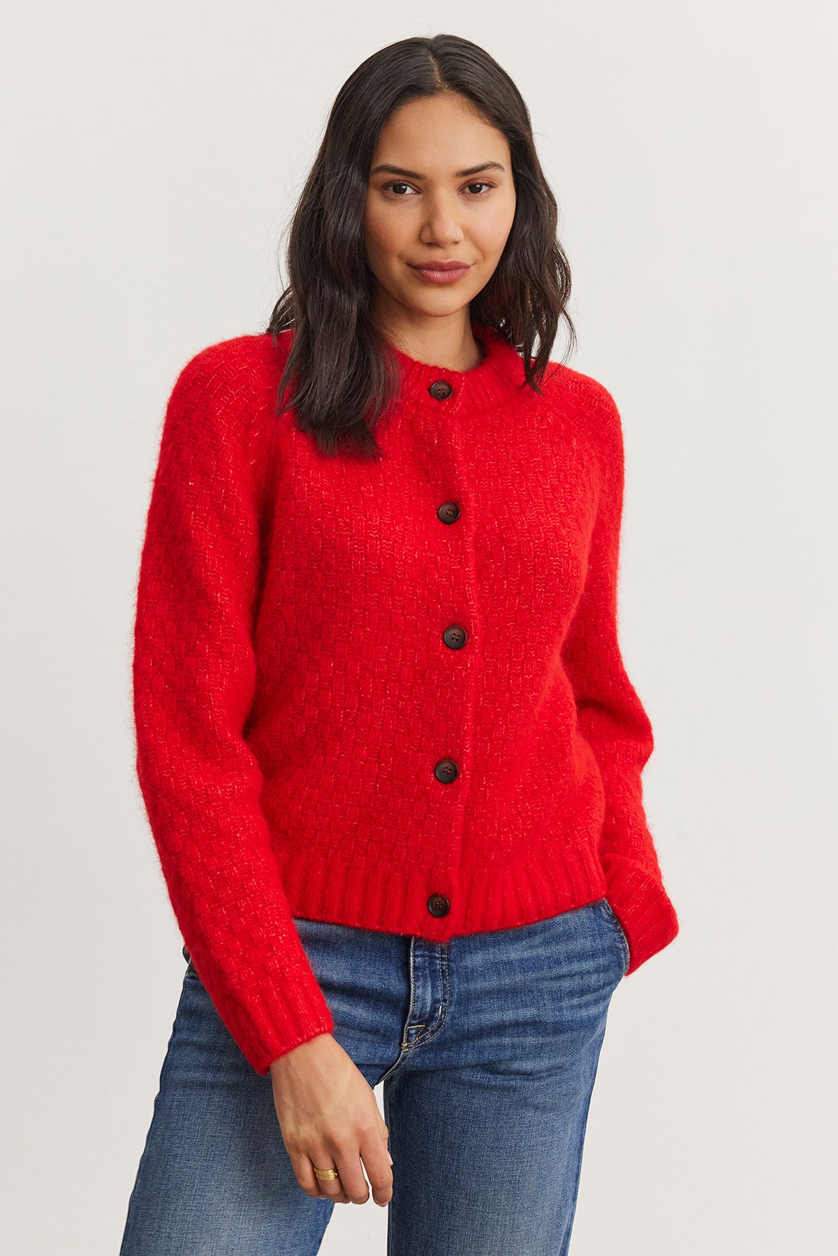   A woman wearing the CHANTAL ALPACA CARDIGAN from Velvet by Graham & Spencer, featuring a red alpaca blend with black buttons, is paired with blue jeans and stands against a light background. 