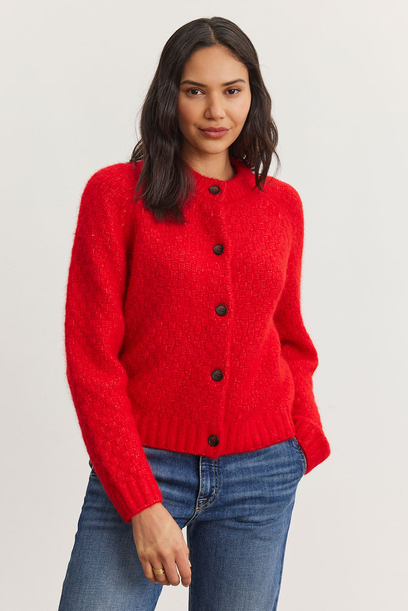 A woman wearing the CHANTAL ALPACA CARDIGAN from Velvet by Graham & Spencer, featuring a red alpaca blend with black buttons, is paired with blue jeans and stands against a light background.