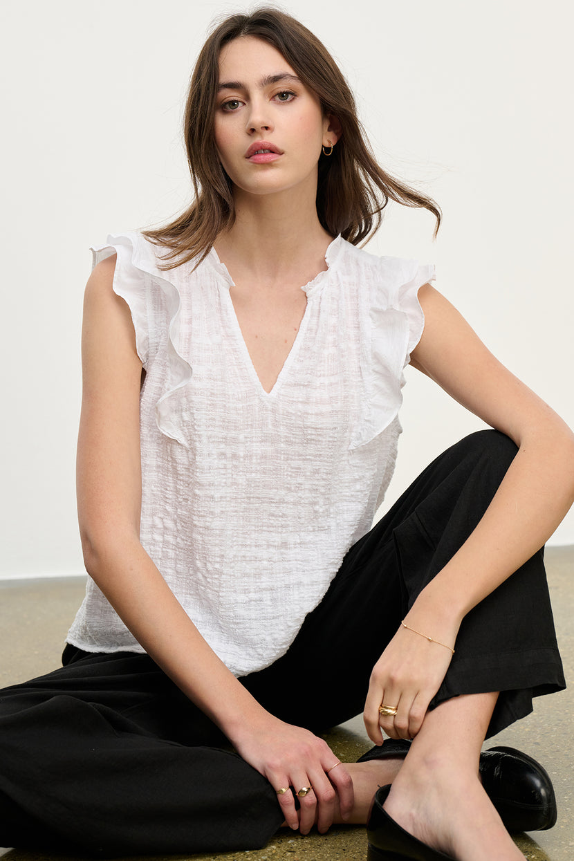 A woman sits gracefully on the floor, wearing the EVA TEXTURED COTTON GAUZE TOP by Velvet by Graham & Spencer, featuring ruffle sleeves and a chic V-neckline, paired with sleek black pants.