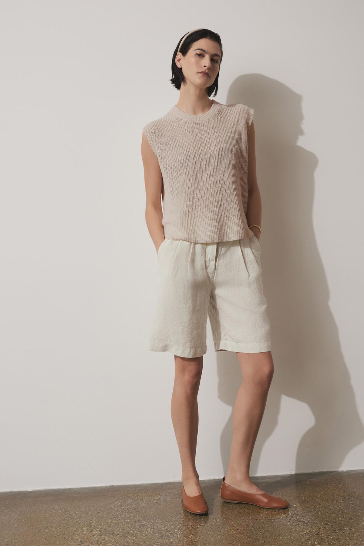   Person standing against a white wall wearing the GARDENA LINEN SWEATER VEST from Velvet by Jenny Graham, light-colored shorts made of breathable linen, a headband, and brown shoes. 