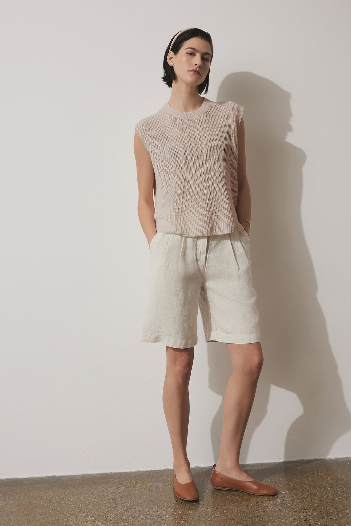   A person stands against a white wall wearing a sleeveless beige top, light-colored LARCHMONT HEAVY LINEN SHORT by Velvet by Jenny Graham, a beige hairband, and brown flat shoes. Hands are in pockets and legs are slightly apart—perfect warm-weather styling. 