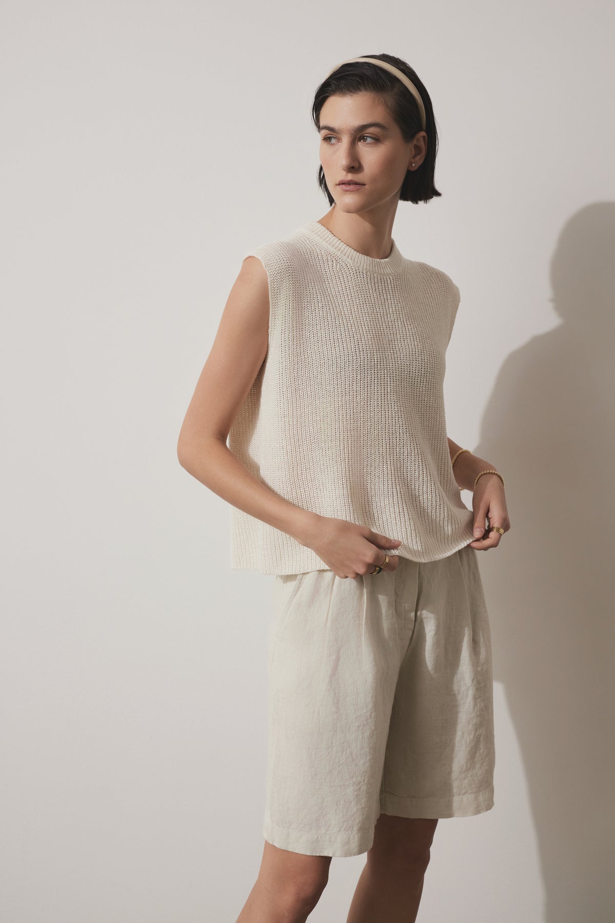   A person with short dark hair is wearing the GARDENA LINEN SWEATER VEST by Velvet by Jenny Graham, paired with relaxed fit shorts made of breathable linen. They are standing against a plain, light background. 