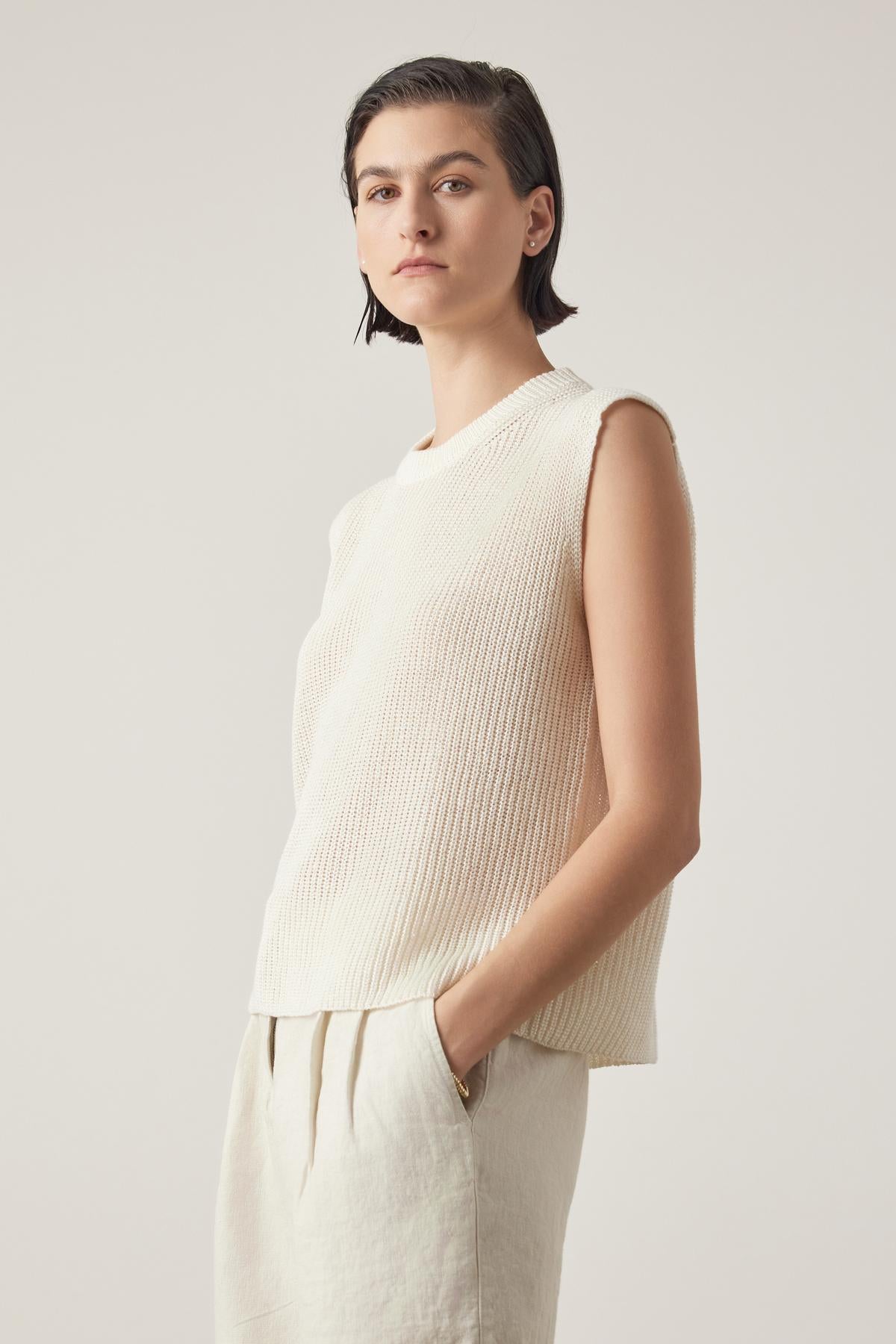   A person with short hair wears the GARDENA LINEN SWEATER VEST by Velvet by Jenny Graham and relaxed fit, light-colored pants. They stand with one hand in their pocket against a plain background. 