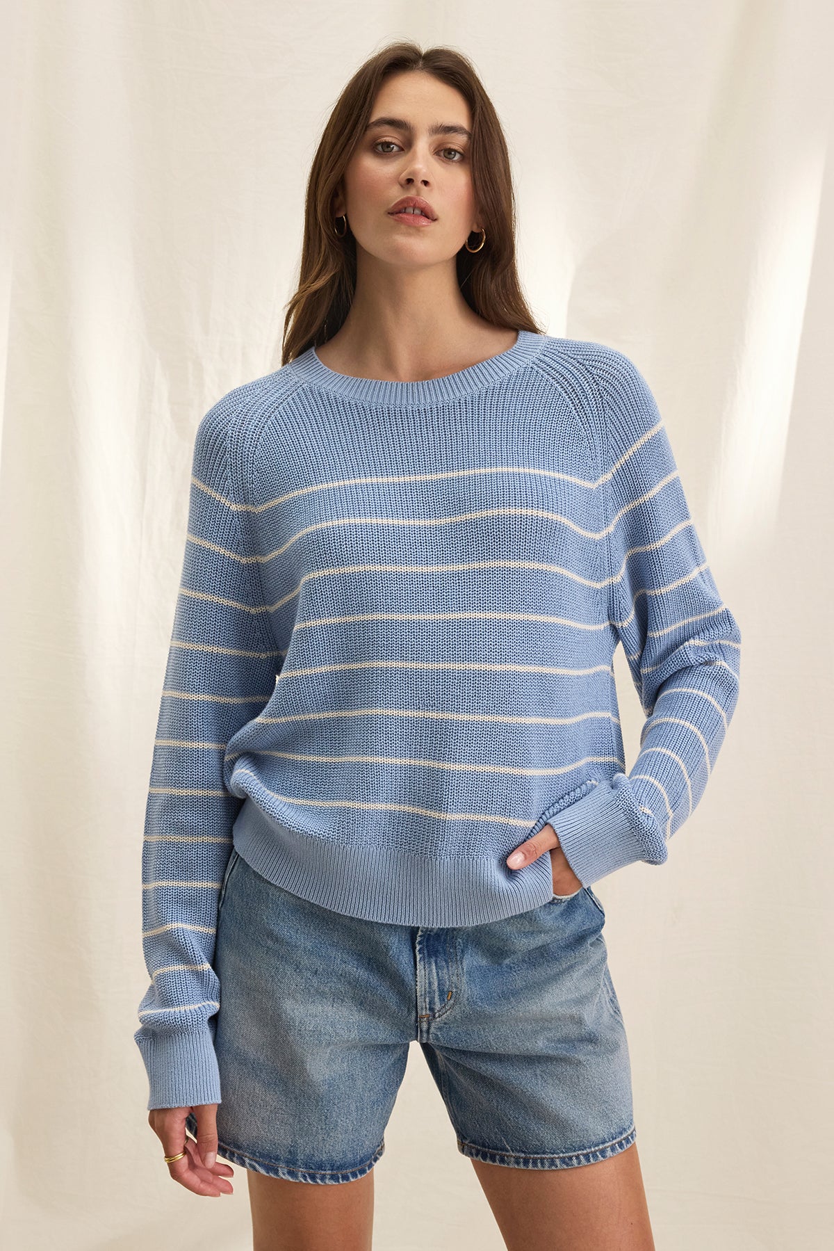   Wearing the Velvet by Graham & Spencer CHAYSE SWEATER with denim shorts epitomizes casual wear, standing out effortlessly against a light, textured background. 