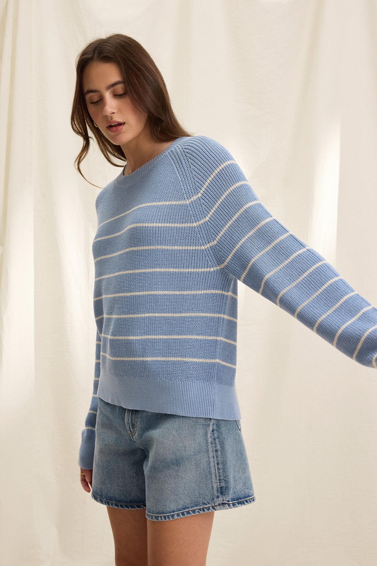 A person in a CHAYSE SWEATER by Velvet by Graham & Spencer, crafted from textured cotton, and denim shorts stands before a light-colored curtain, showcasing a relaxed silhouette ideal for casual wear.-38662654853313