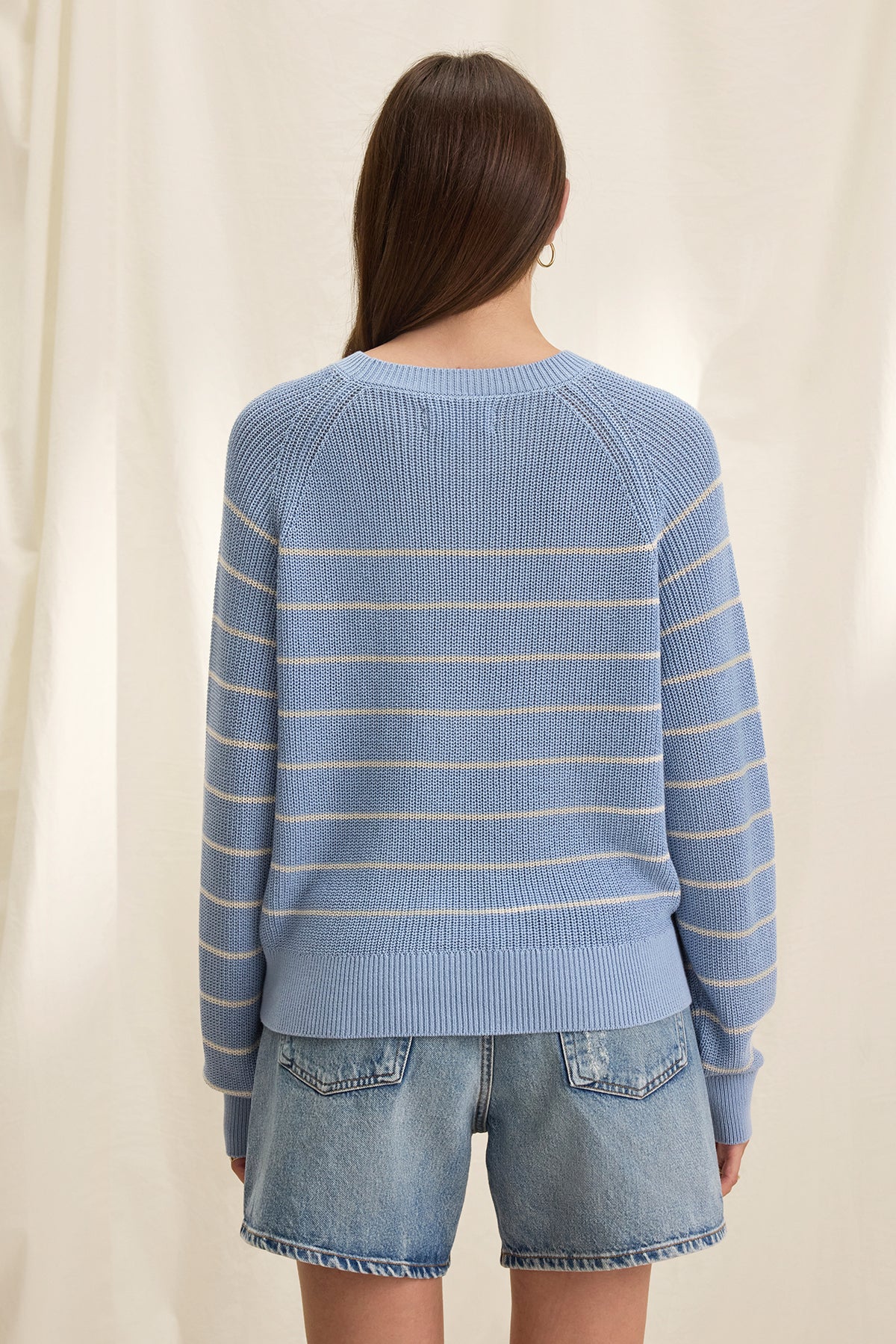 Back view of a person with long hair wearing the CHAYSE SWEATER by Velvet by Graham & Spencer, featuring a blue and white textured cotton stripe design and paired with denim shorts, creating a relaxed silhouette against a light background.-38662654886081