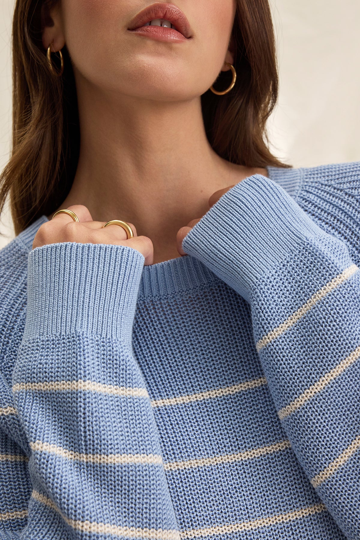 The individual is wearing the CHAYSE SWEATER from Velvet by Graham & Spencer, a light blue knit with white stripes and a relaxed fit. Their light brown hair highlights gold hoop earrings and rings. The textured cotton sweater's sleeves drape slightly over their hands, ideal for casual wear.-38662654918849