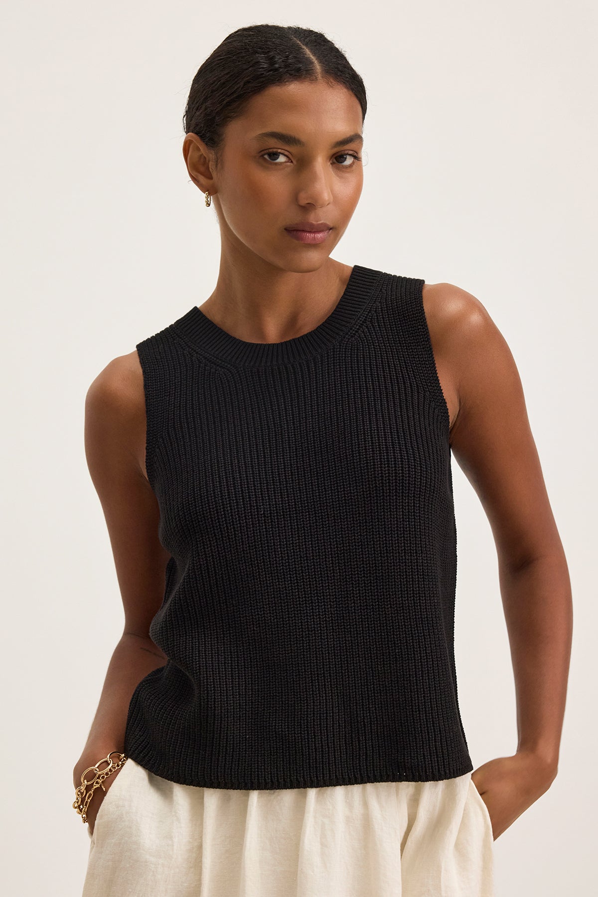 A woman stands against a plain background wearing the MUNA SWEATER VEST by Velvet by Graham & Spencer, a sleeveless black knit top ideal for layering, paired with beige pants.-38662690930881