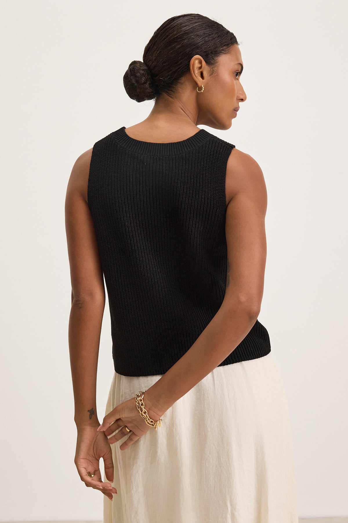   With short, dark hair styled in a bun, the individual exudes a modern touch wearing the MUNA SWEATER VEST by Velvet by Graham & Spencer paired with textured cotton white pants, standing with hands crossed behind their back and facing slightly away from the camera. 