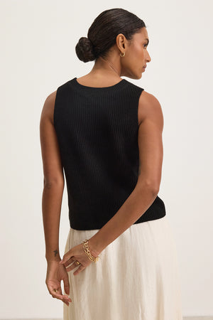 With short, dark hair styled in a bun, the individual exudes a modern touch wearing the MUNA SWEATER VEST by Velvet by Graham & Spencer paired with textured cotton white pants, standing with hands crossed behind their back and facing slightly away from the camera.