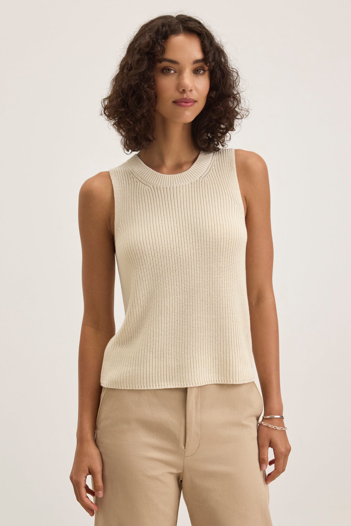   A person with curly hair wears a chic Velvet by Graham & Spencer MUNA SWEATER VEST in beige, crafted from textured cotton, paired with matching pants, against a plain background. 