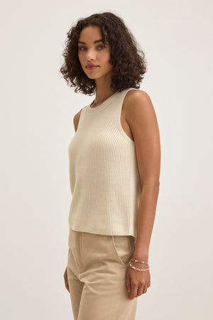 A person with curly hair wears the MUNA SWEATER VEST by Velvet by Graham & Spencer, a beige textured cotton sleeveless top, paired with light pants, standing against a plain background.