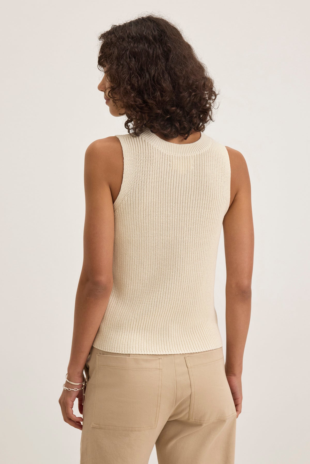   A person with curly hair wears the MUNA SWEATER VEST by Velvet by Graham & Spencer along with beige pants, viewed from behind against a plain backdrop. 