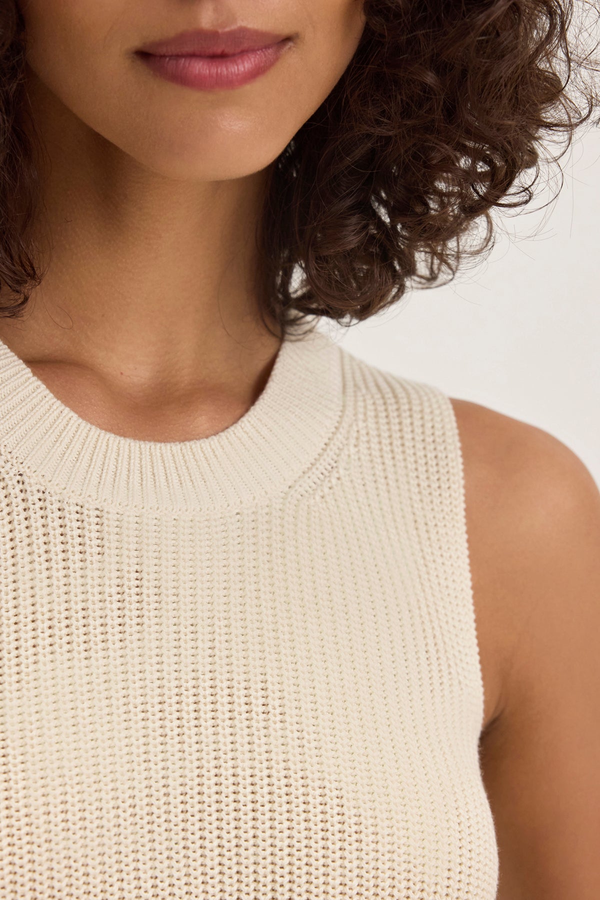   A person with curly hair wears the MUNA SWEATER VEST by Velvet by Graham & Spencer. This sleeveless, textured cotton top enhances the upper torso and neckline, blending style with comfort. 