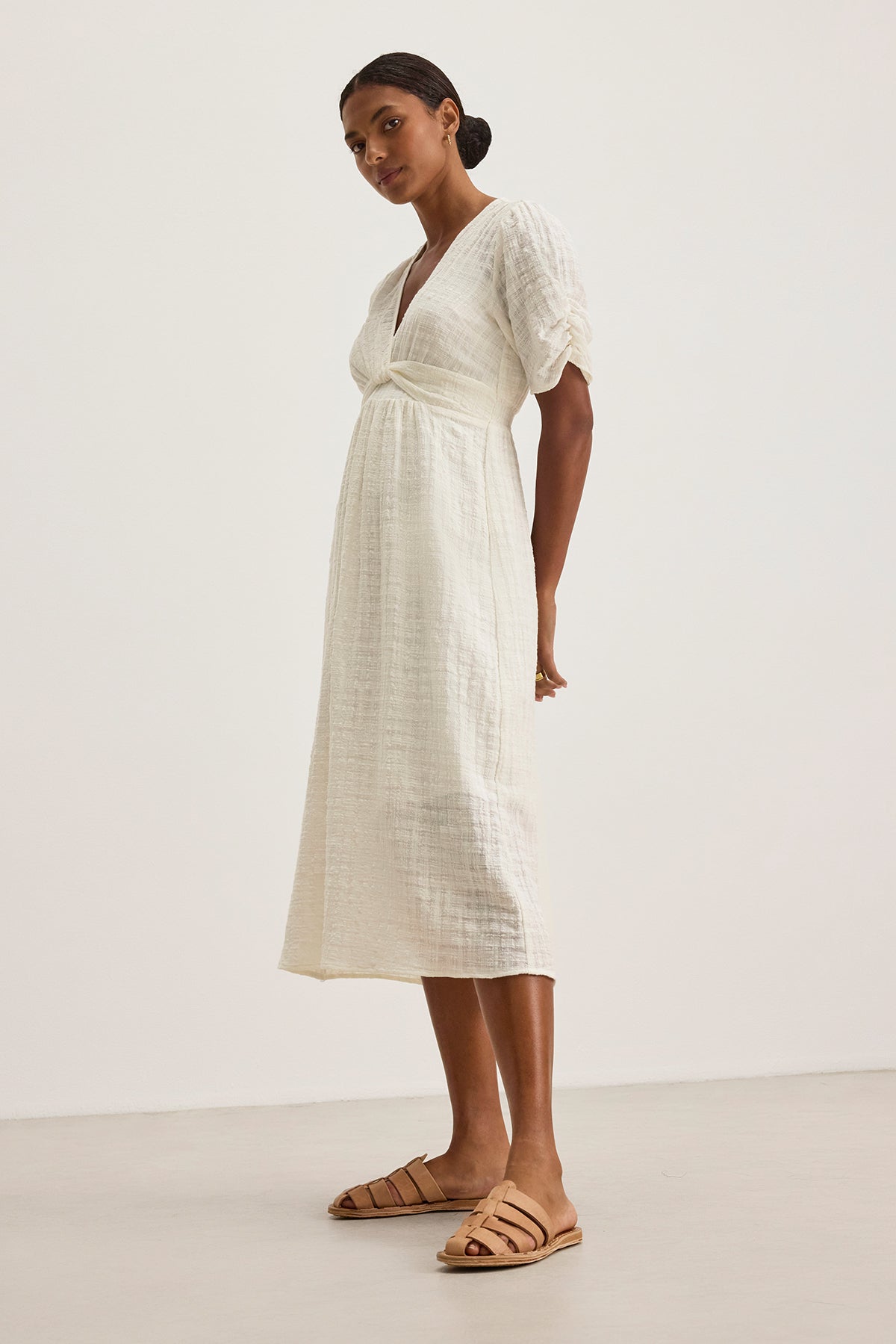   A person in the ELOISE TEXTURED COTTON GAUZE DRESS by Velvet by Graham & Spencer stands sideways on a plain background, wearing tan sandals. The white midi dress features a v-neckline and short sleeves. 