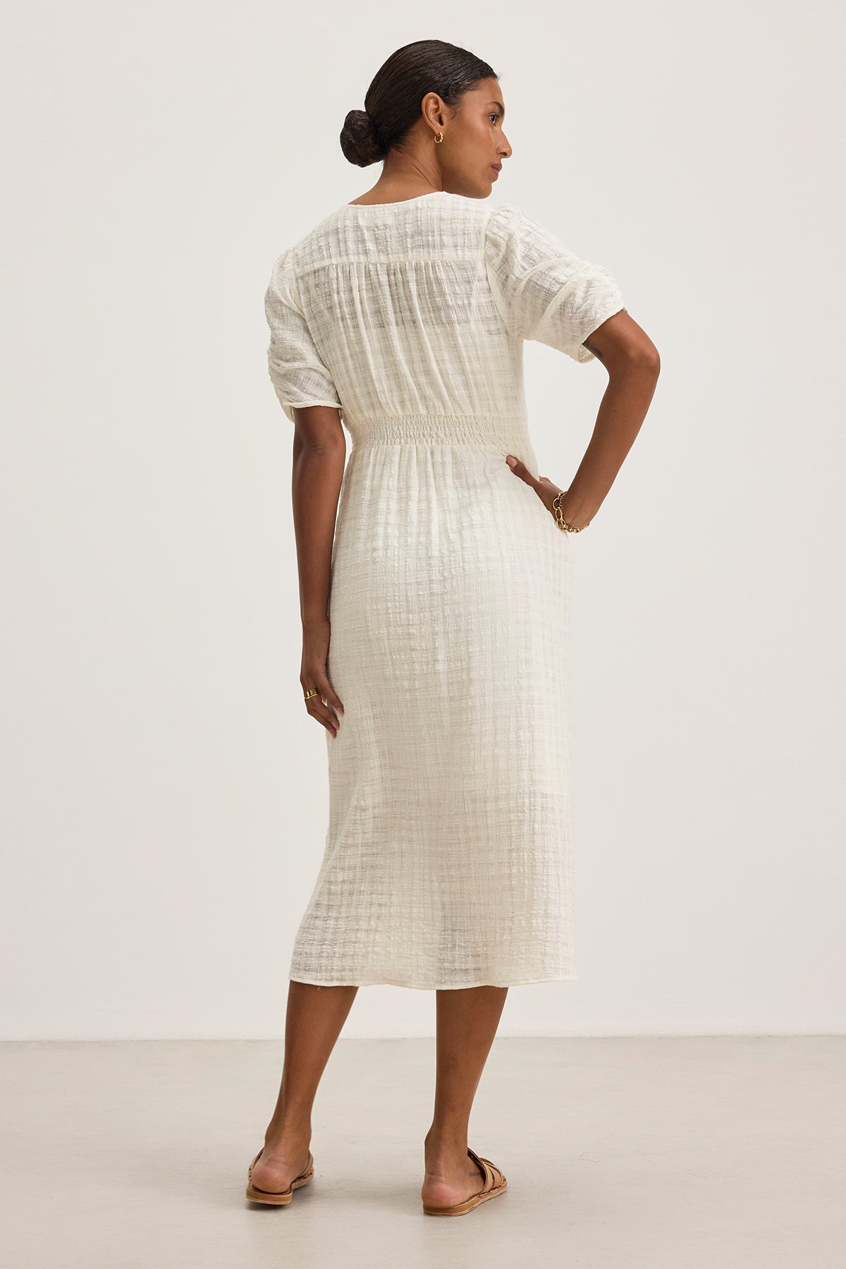   Against a plain background, a woman stands facing away, wearing the ELOISE TEXTURED COTTON GAUZE DRESS by Velvet by Graham & Spencer. The dress is white with a v-neckline and paired with sandals. 