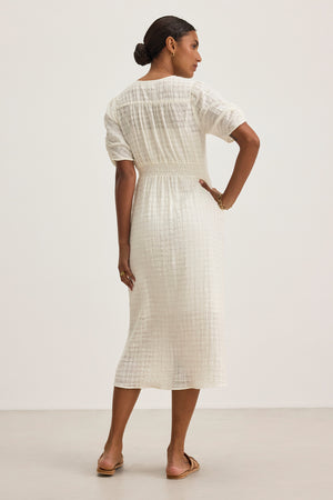 Against a plain background, a woman stands facing away, wearing the ELOISE TEXTURED COTTON GAUZE DRESS by Velvet by Graham & Spencer. The dress is white with a v-neckline and paired with sandals.