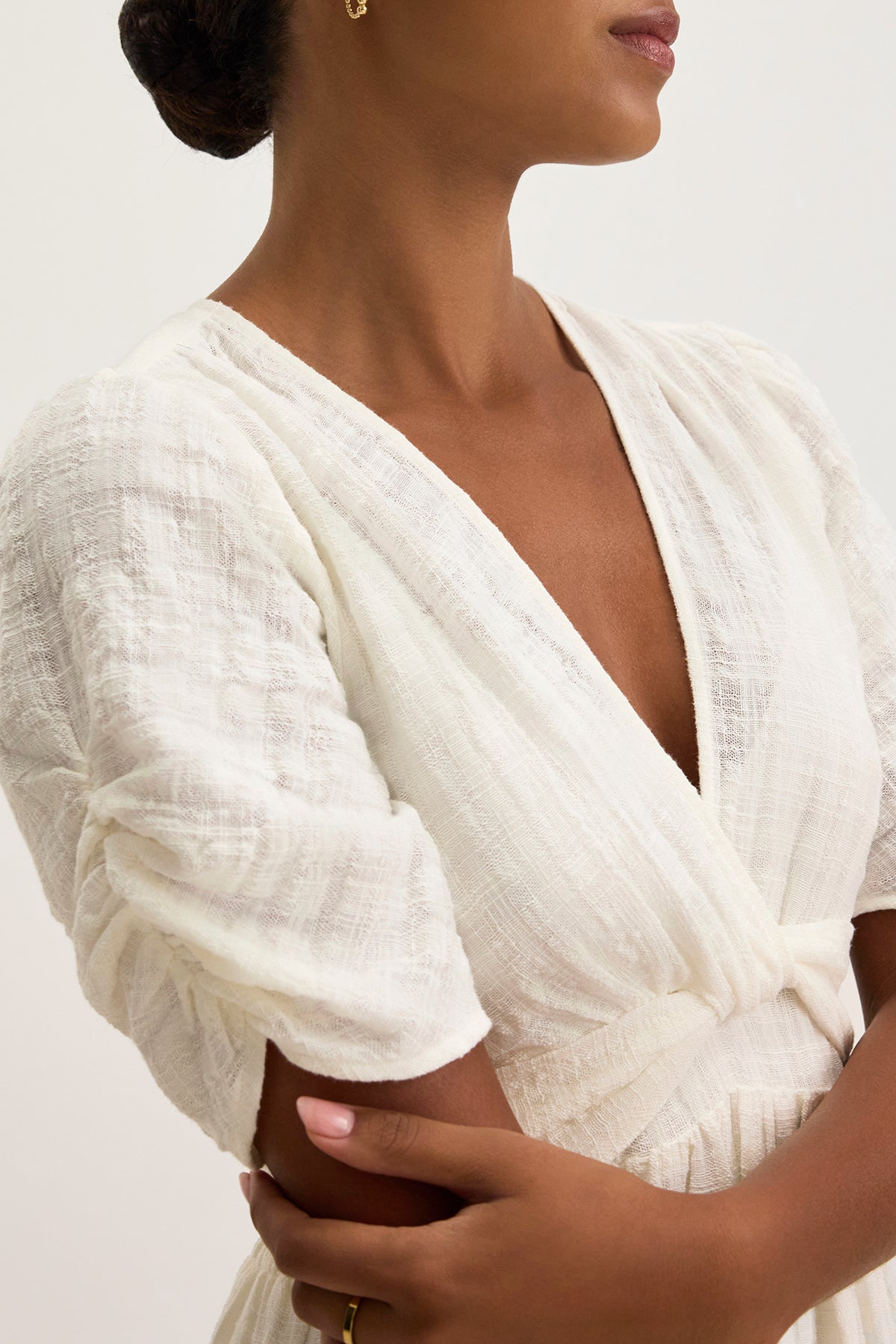   A person in the ELOISE TEXTURED COTTON GAUZE DRESS by Velvet by Graham & Spencer, featuring a white midi wrap style with short pouf sleeves and a v-neckline accentuated with a gold ring, stands against a neutral background. 