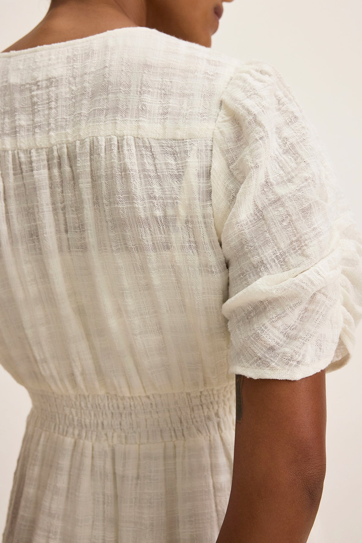   A person elegantly models the ELOISE TEXTURED COTTON GAUZE DRESS by Velvet by Graham & Spencer. This white midi dress features puffed sleeves, an elastic cinched waist, and a subtle v-neckline as they pose against a simple backdrop. 