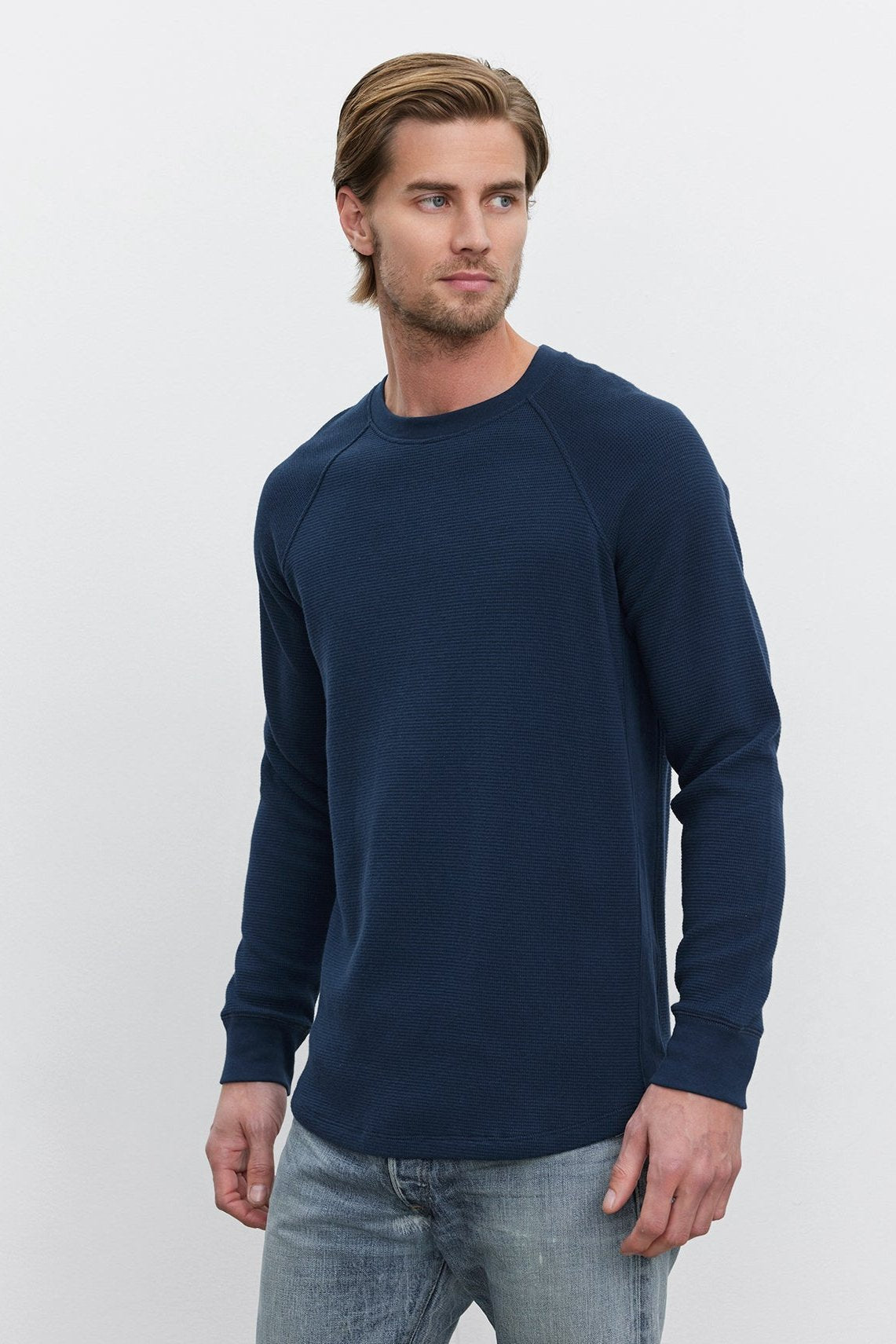   A man with light brown hair is wearing the GLEN THERMAL CREW by Velvet by Graham & Spencer in navy blue, paired with light blue jeans, and standing against a plain white background. 