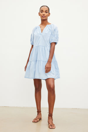 A person stands against a plain background wearing the CHRISSY DRESS by Velvet by Graham & Spencer, which is a light blue, short-sleeve, knee-length cotton dress with floral embroidery. The dress features a V-neckline and tiered skirt and is paired with brown strappy sandals.