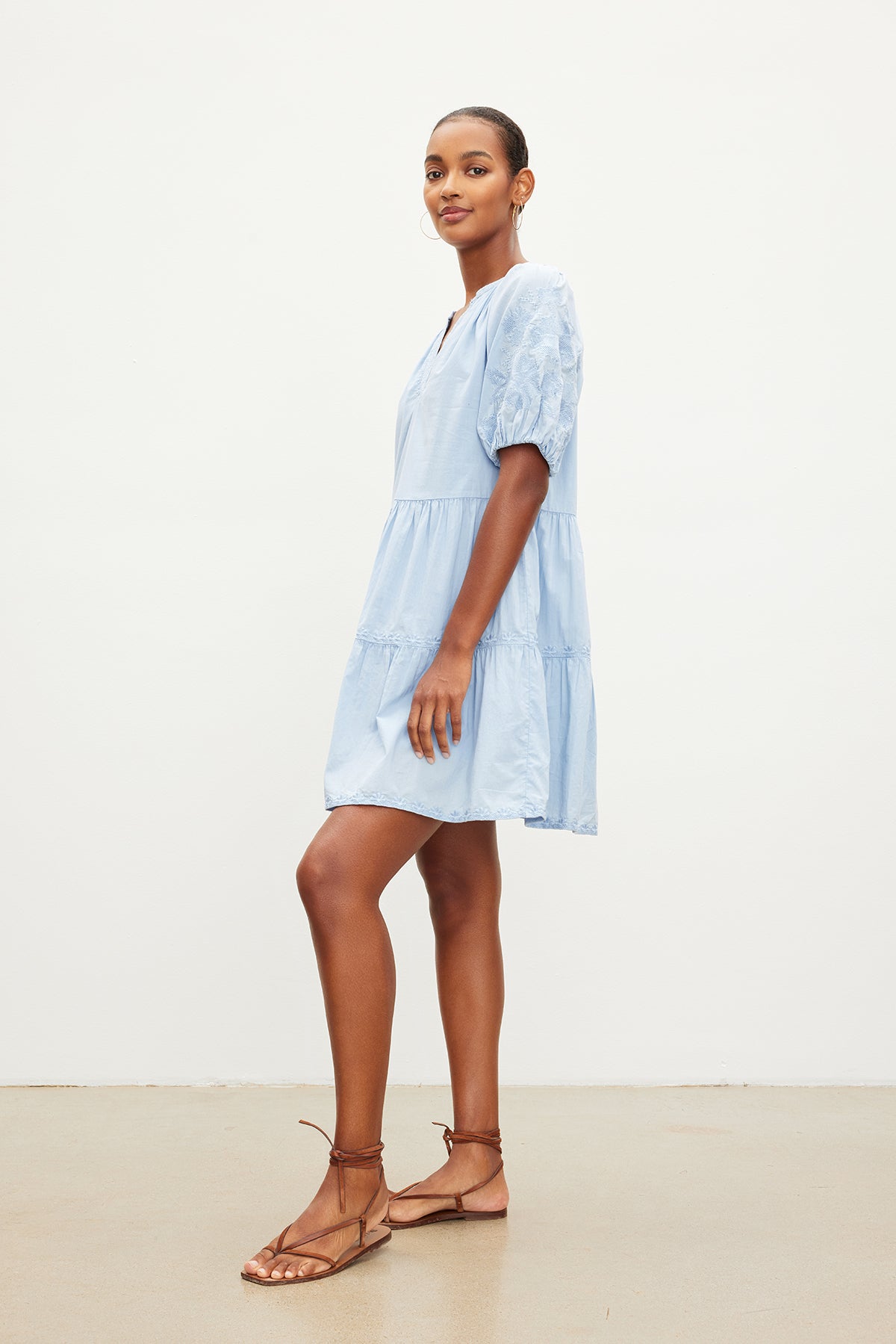   Person in the CHRISSY DRESS by Velvet by Graham & Spencer, a light blue, short-sleeved, knee-length cotton dress with floral embroidery, stands sideways while wearing brown sandals. 