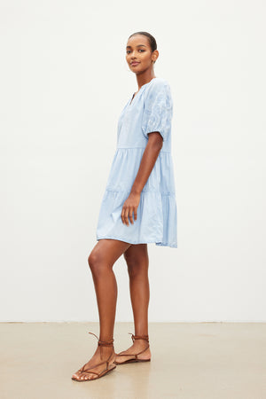 Person in the CHRISSY DRESS by Velvet by Graham & Spencer, a light blue, short-sleeved, knee-length cotton dress with floral embroidery, stands sideways while wearing brown sandals.