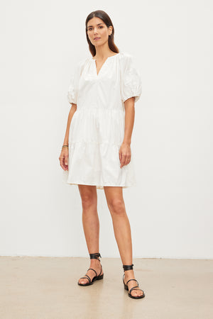 A person stands against a plain background, wearing the CHRISSY DRESS from Velvet by Graham & Spencer, a white, short-sleeve, knee-length cotton dress with floral embroidery and black sandals.