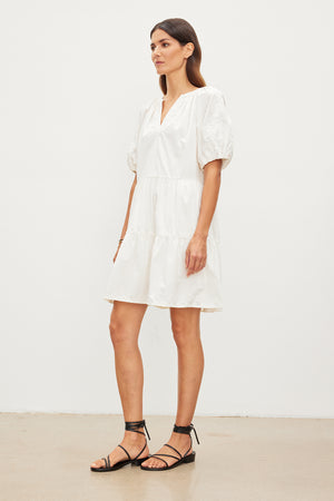 A woman wearing the CHRISSY DRESS by Velvet by Graham & Spencer, characterized by its white color, short sleeves, and v-neckline, stands against a plain white background while sporting black strappy sandals.