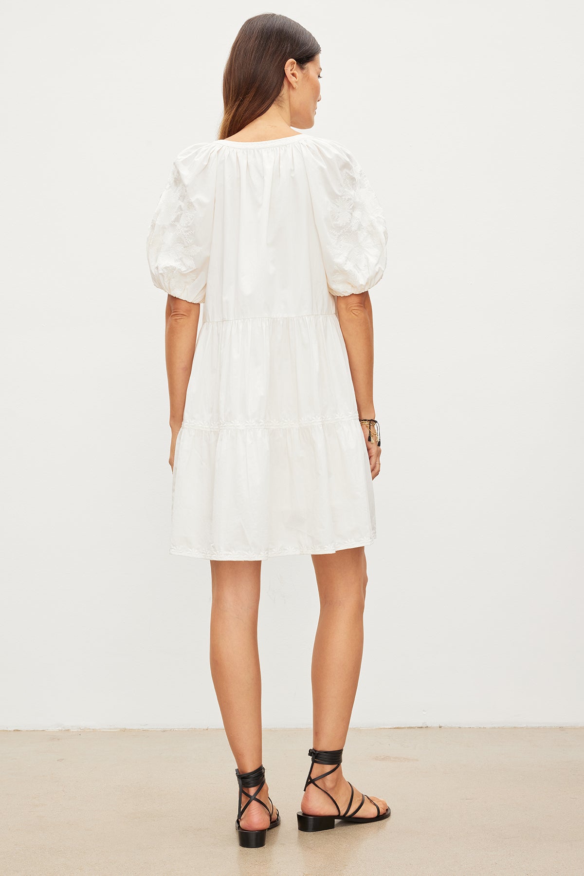   A woman with long brown hair, facing away, is wearing a Velvet by Graham & Spencer CHRISSY DRESS, a white knee-length cotton dress with puff sleeves and black sandals. She stands in front of a plain white background. The dress features delicate floral embroidery that adds a touch of elegance. 