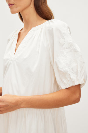 A person wearing the CHRISSY DRESS by Velvet by Graham & Spencer, a white blouse with a v-neckline and short puff sleeves adorned with floral embroidery, stands in a neutral setting.