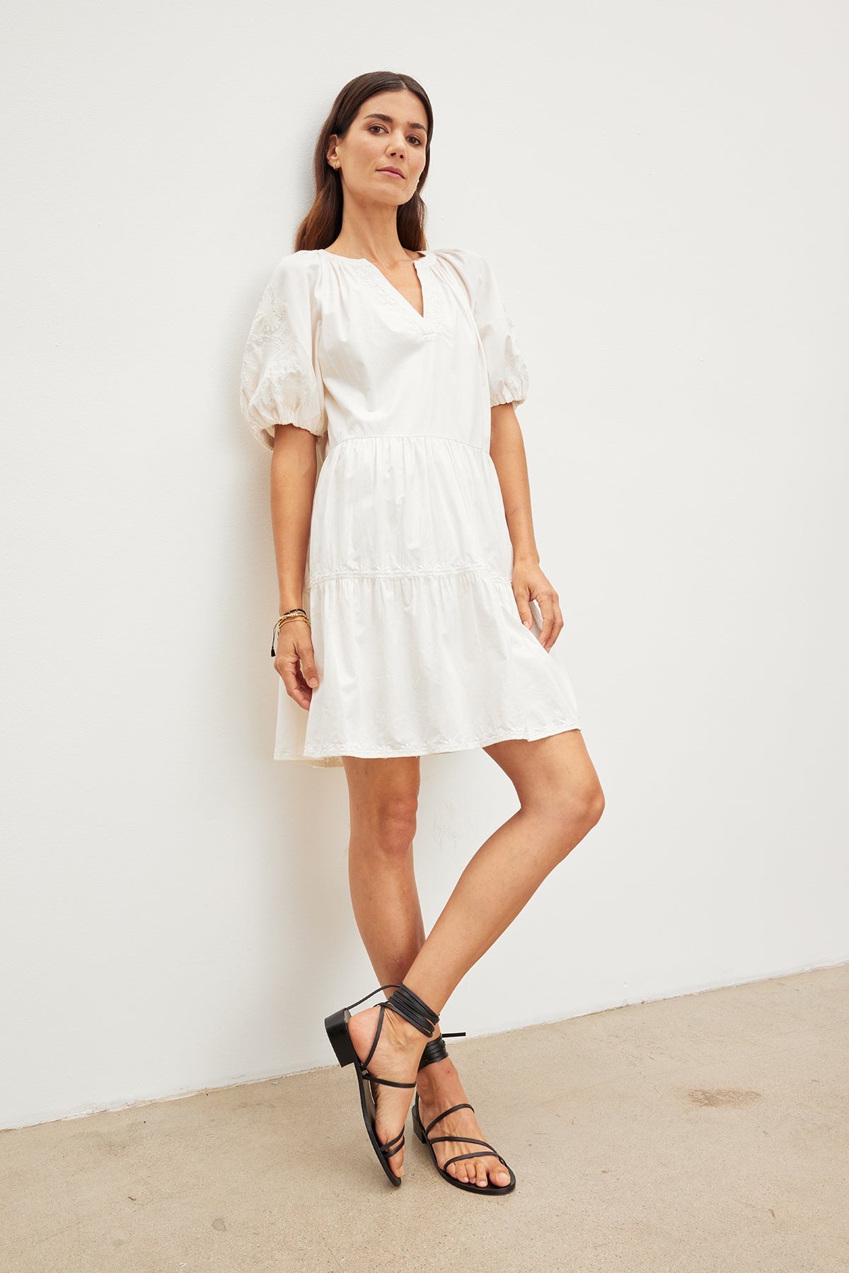   A woman stands against a plain white wall wearing the CHRISSY DRESS by Velvet by Graham & Spencer, a short white cotton dress with puffy sleeves and a v-neckline, subtly adorned with floral embroidery. She pairs it with black strappy sandals. She has long, straight hair and a relaxed expression. 