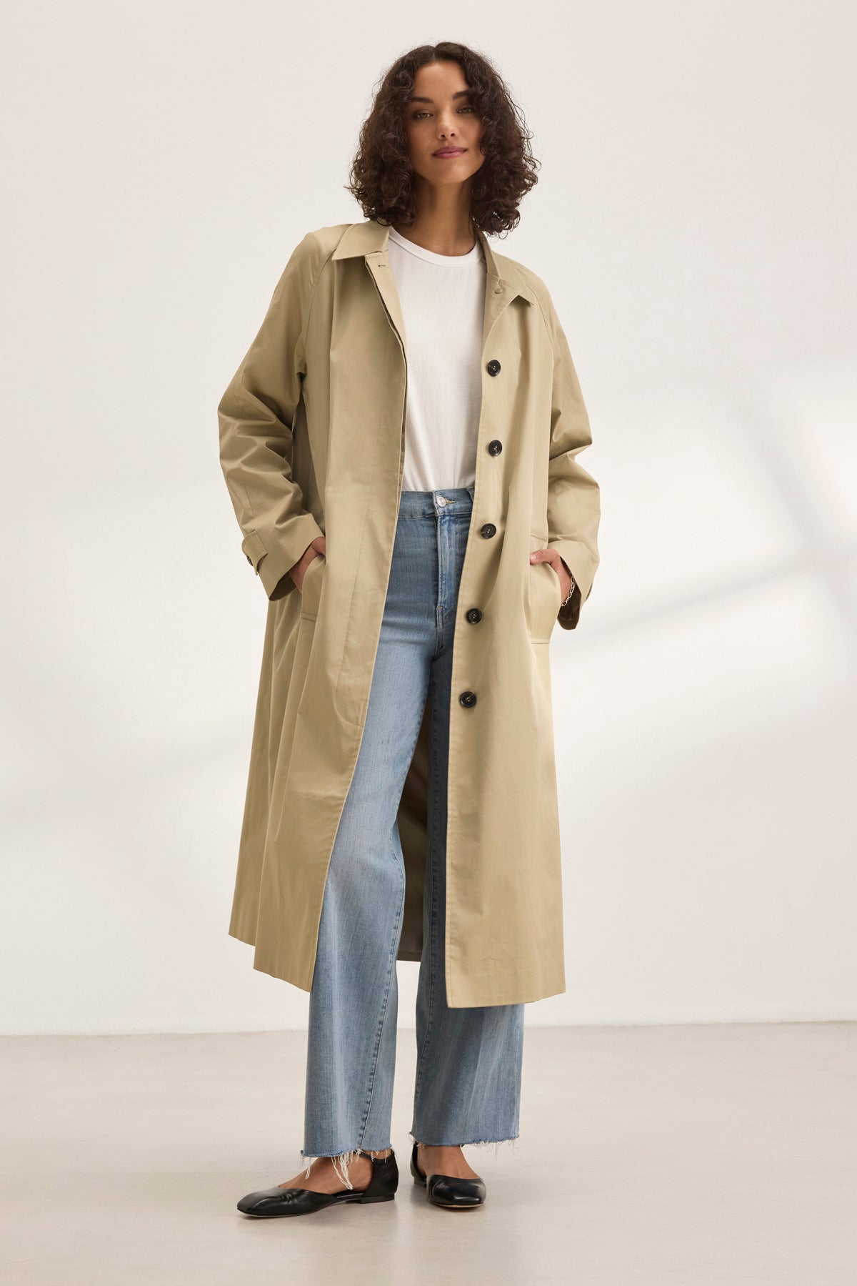   Someone wears the RAHEAL TRENCH COAT by Velvet by Graham & Spencer, featuring a cotton-elastane shell in beige. Layered over a white shirt and jeans, they stand in a well-lit room with hands casually in pockets, showcasing versatile styling. 