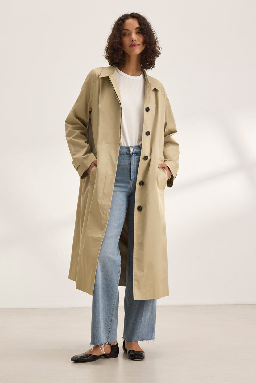 Someone wears the RAHEAL TRENCH COAT by Velvet by Graham & Spencer, featuring a cotton-elastane shell in beige. Layered over a white shirt and jeans, they stand in a well-lit room with hands casually in pockets, showcasing versatile styling.