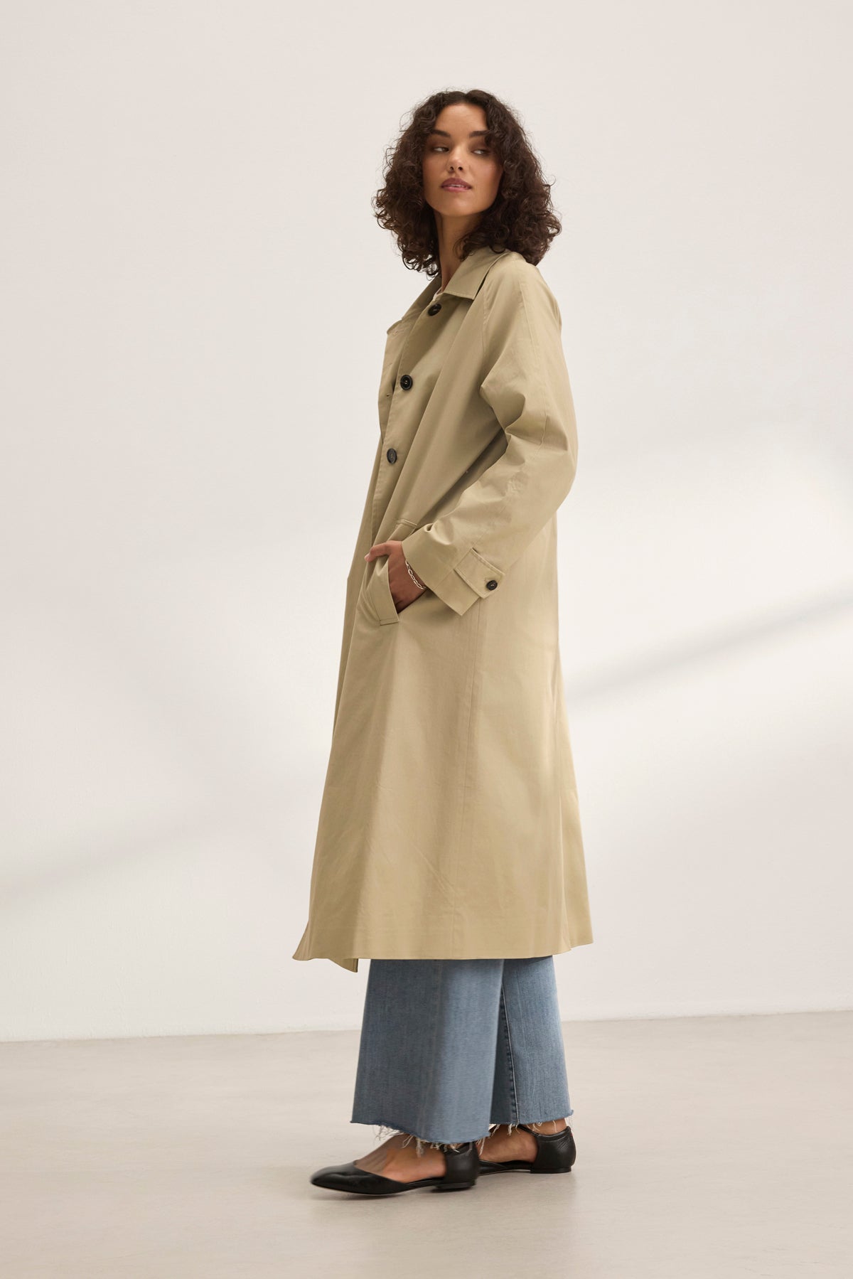   Against a plain background, a person exudes elegance in the RAHEAL TRENCH COAT by Velvet by Graham & Spencer, paired with blue jeans and black shoes. With hands casually in pockets, this classic beige trench enhances any style effortlessly. 