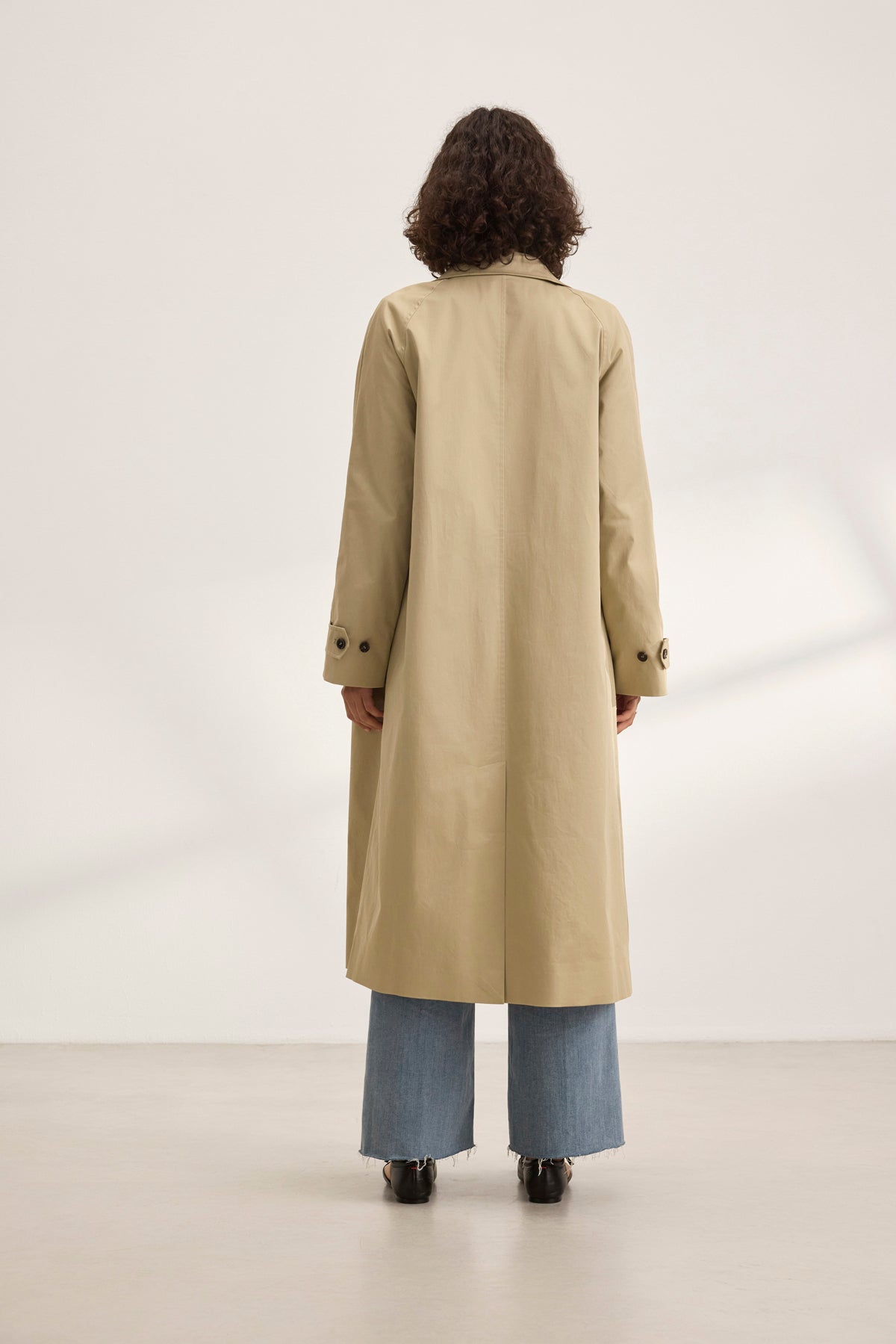   In a minimalist white room, a person with curly hair models the RAHEAL TRENCH COAT by Velvet by Graham & Spencer and blue jeans, epitomizing versatile styling. 