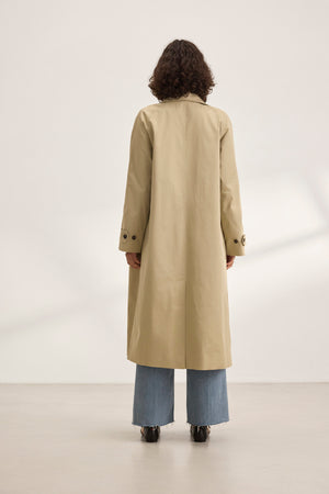 In a minimalist white room, a person with curly hair models the RAHEAL TRENCH COAT by Velvet by Graham & Spencer and blue jeans, epitomizing versatile styling.