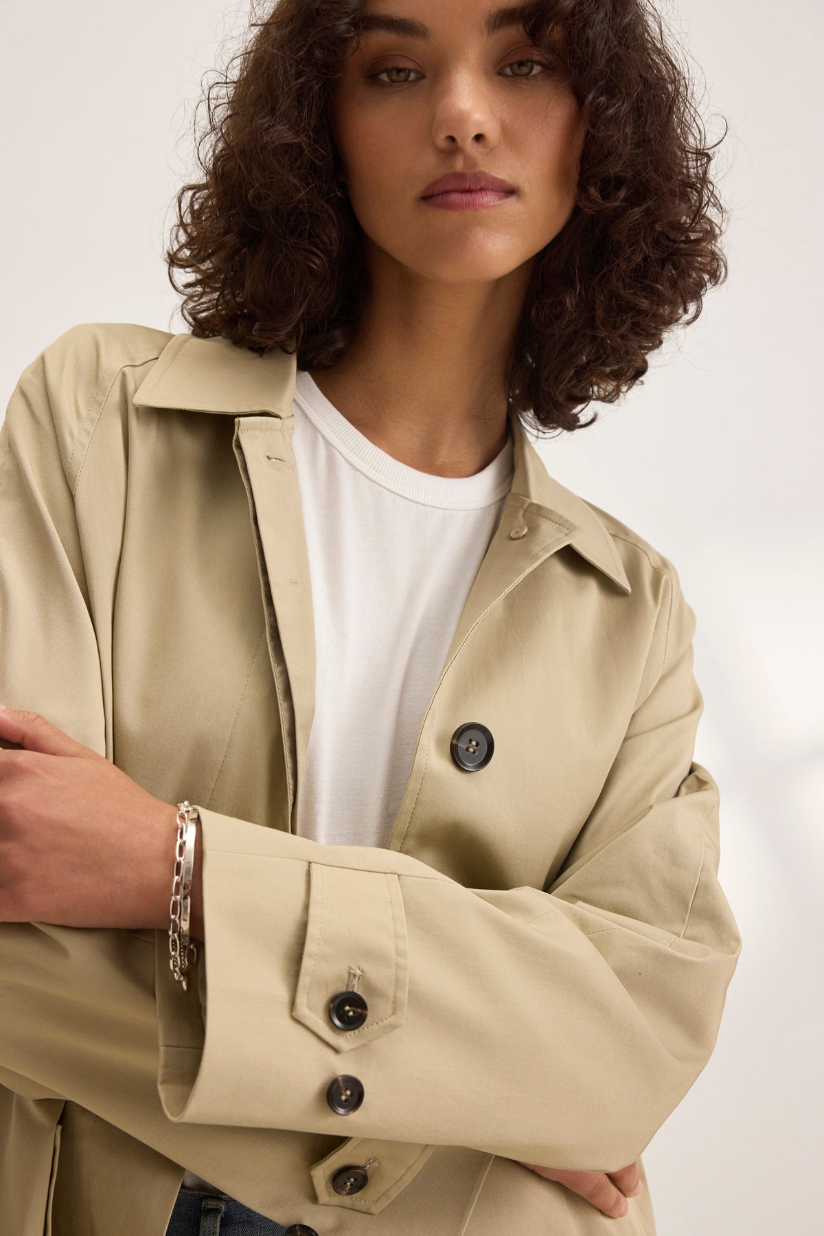   The curly-haired individual stands confidently, arms crossed, wearing the RAHEAL TRENCH COAT by Velvet by Graham & Spencer. The beige coat features a detachable belt over their crisp white shirt. 