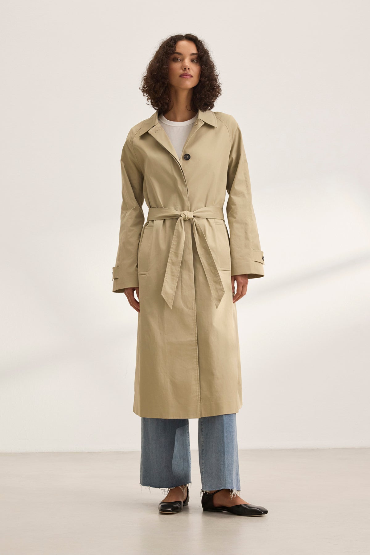 A person models the versatile RAHEAL TRENCH COAT by Velvet by Graham & Spencer. They pair it with light blue jeans and black shoes against a plain backdrop, highlighting the beige cotton-elastane coat's stylish design with a belt.-38819765780673