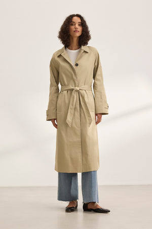 A person models the versatile RAHEAL TRENCH COAT by Velvet by Graham & Spencer. They pair it with light blue jeans and black shoes against a plain backdrop, highlighting the beige cotton-elastane coat's stylish design with a belt.