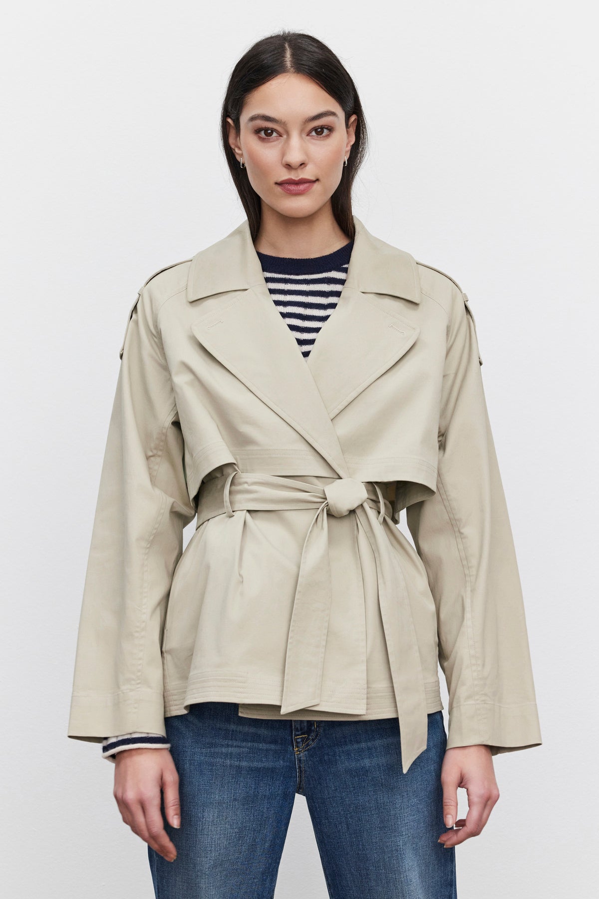   A person with long dark hair stands against a plain backdrop, wearing the NANCY TRENCH JACKET by Velvet by Graham & Spencer, complete with a detachable belt, over a black-and-white striped top and blue jeans. 