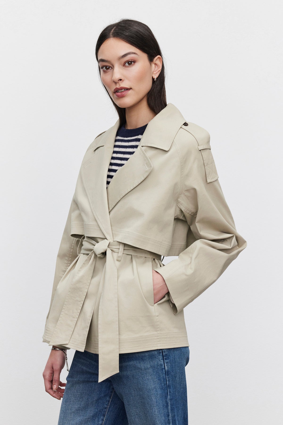 A person with long dark hair wearing the NANCY TRENCH JACKET by Velvet by Graham & Spencer over a striped shirt and jeans, standing with one hand in their pocket.-37676720750785