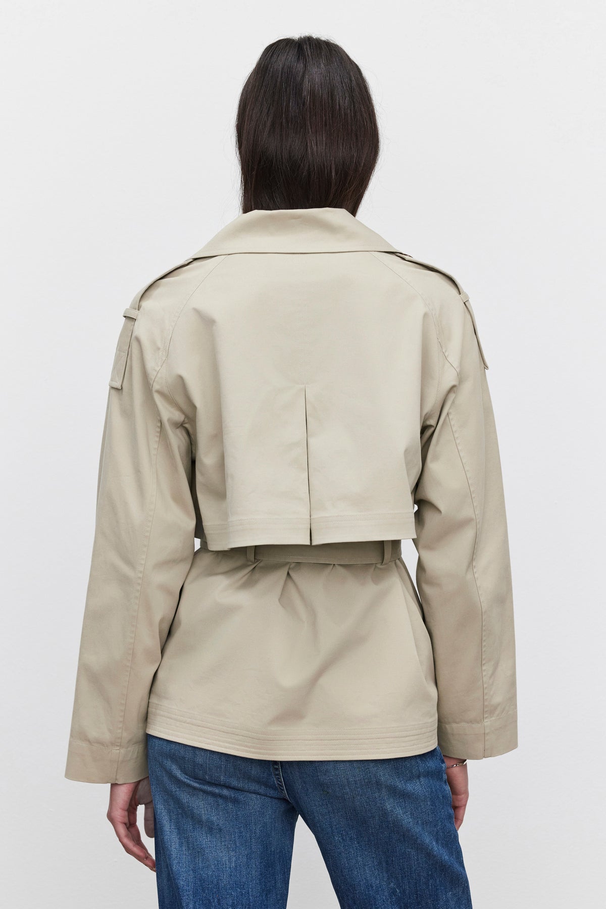   A person wearing the NANCY TRENCH JACKET by Velvet by Graham & Spencer, along with blue jeans, is shown from the back against a plain white background. The tan, waist-length jacket with long sleeves resembles a modern trench coat style and includes a detachable belt for added versatility. 