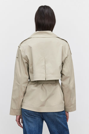 A person wearing the NANCY TRENCH JACKET by Velvet by Graham & Spencer, along with blue jeans, is shown from the back against a plain white background. The tan, waist-length jacket with long sleeves resembles a modern trench coat style and includes a detachable belt for added versatility.