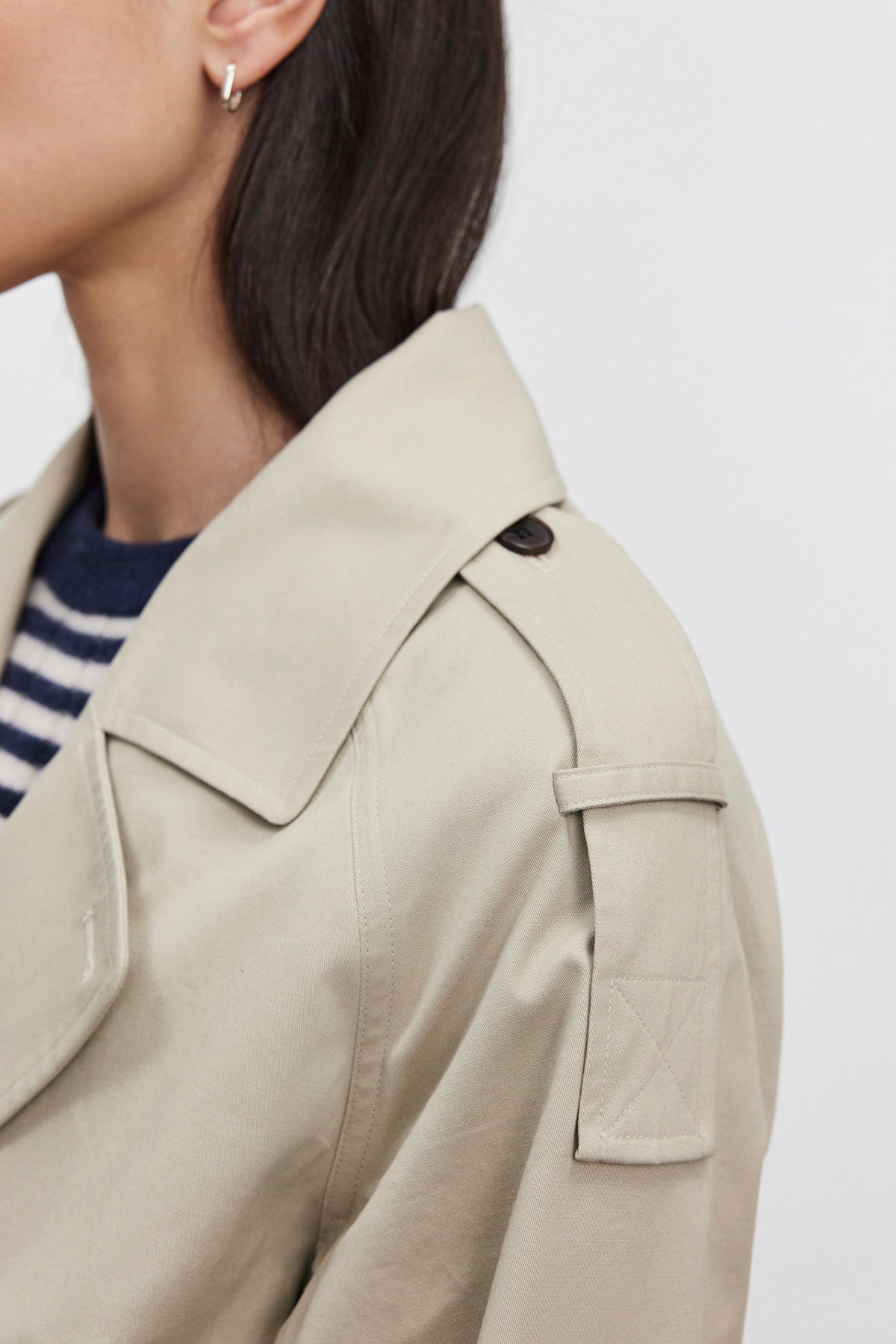   Close-up of a person in the NANCY TRENCH JACKET by Velvet by Graham & Spencer, crafted from cotton elastane and featuring a detachable belt, shoulder strap, and button. The individual is also seen wearing a striped shirt and small hoop earring, with only the side of their face and shoulder visible. 