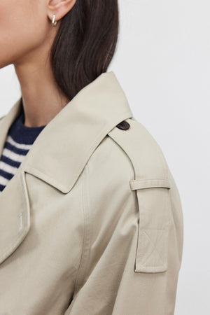 Close-up of a person in the NANCY TRENCH JACKET by Velvet by Graham & Spencer, crafted from cotton elastane and featuring a detachable belt, shoulder strap, and button. The individual is also seen wearing a striped shirt and small hoop earring, with only the side of their face and shoulder visible.