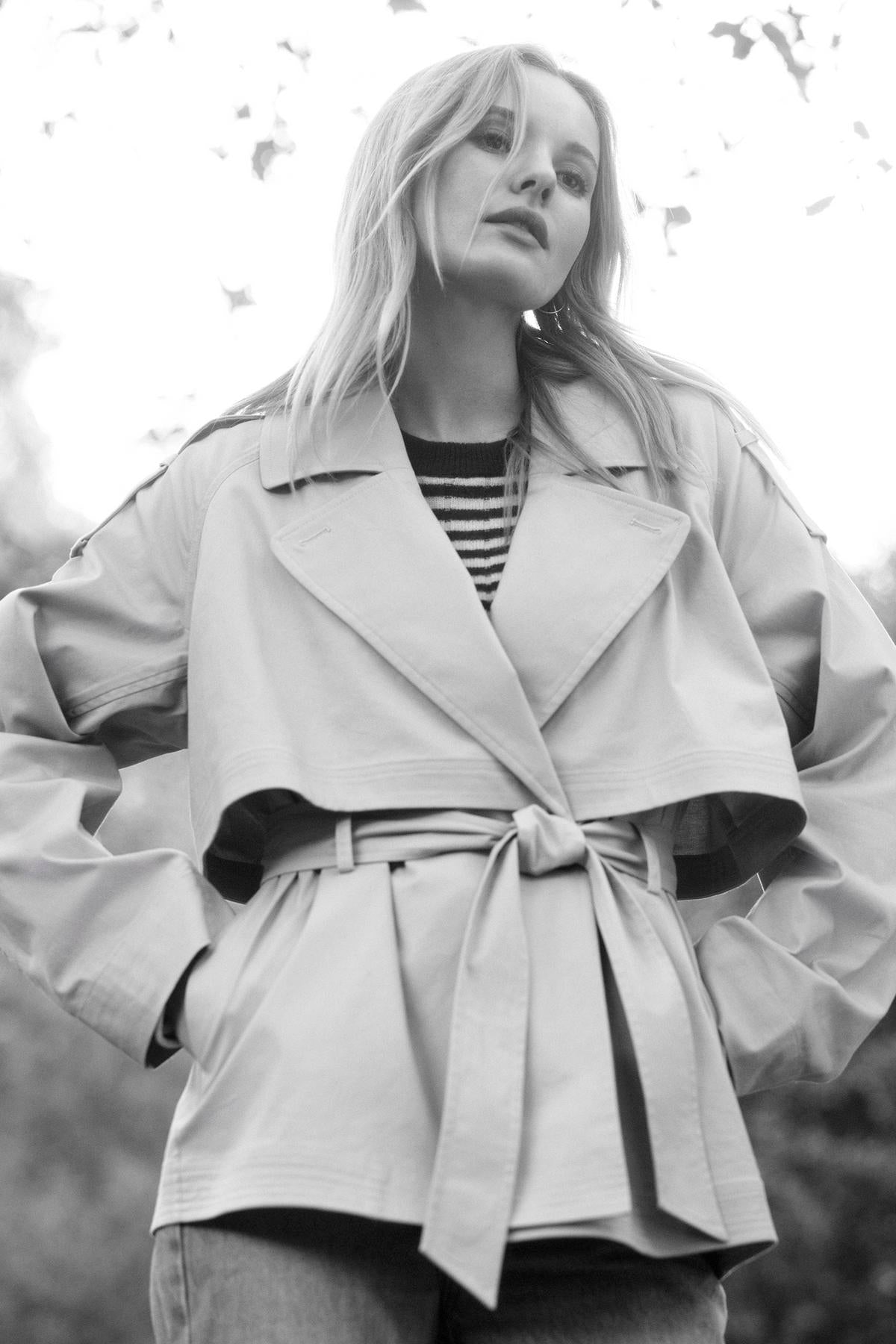 Black and white photo of a woman wearing the NANCY TRENCH JACKET by Velvet by Graham & Spencer, featuring a detachable belt and paired with a striped shirt, standing outdoors with hands in pockets, looking off to the side.-37676720783553