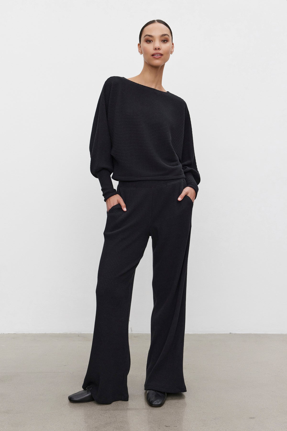   A person stands against a plain background wearing the COLLEEN RIBBED PANT and a matching black long-sleeve top from Velvet by Graham & Spencer, both made from ultra-soft fabric, with hands in pockets. 